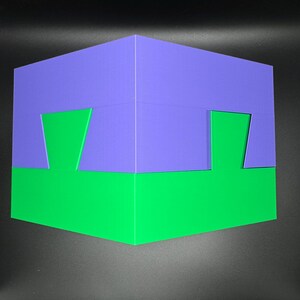 May include: A 3D puzzle box with a purple top and a green bottom. The box has a unique design with cutouts on each side.