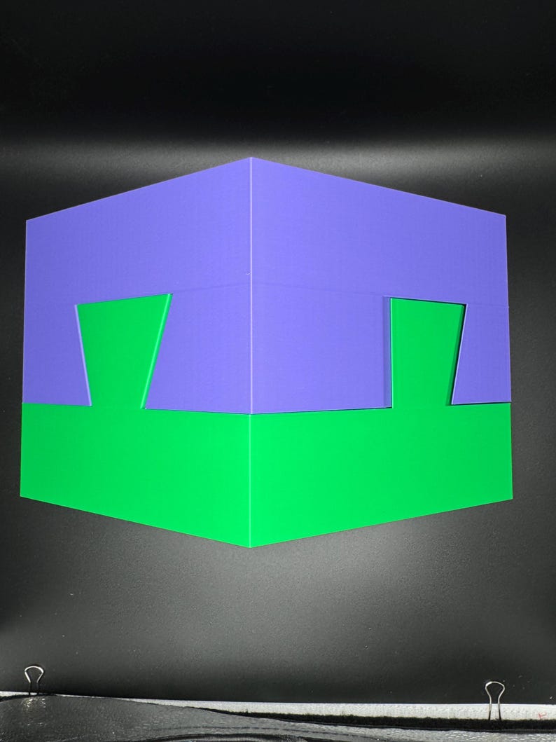 May include: A 3D puzzle box with a purple top and a green bottom. The box has a unique design with cutouts on each side.