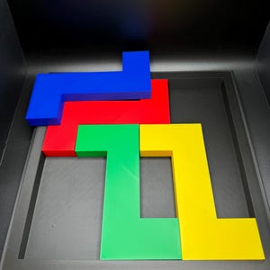 Op de afbeelding: Een kleurrijke puzzel met vier L-vormige stukken in blauw, rood, groen en geel. De stukken zijn gerangschikt in een zwarte bak, wat een visuele uitdaging cre&euml;ert. De levendige kleuren en geometrische vormen maken het een boeiende hersenkraker.