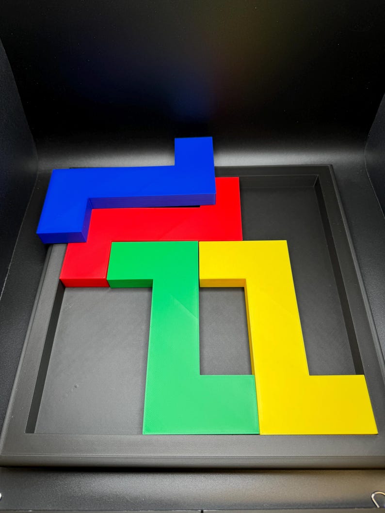 Op de afbeelding: Een kleurrijke puzzel met vier L-vormige stukken in blauw, rood, groen en geel. De stukken zijn gerangschikt in een zwarte bak, wat een visuele uitdaging cre&euml;ert. De levendige kleuren en geometrische vormen maken het een boeiende hersenkraker.
