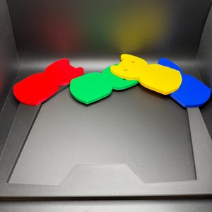 May include: Four colorful, abstract cat-shaped acrylic coasters in red, green, yellow, and blue. The coasters are arranged in a row, with each one slightly overlapping the next. The background is black.