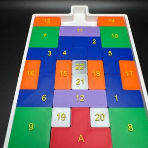 Op de afbeelding: Een witte plastic bak met een puzzelspel. De puzzelstukken zijn gemaakt van verschillende kleuren, waaronder oranje, groen, blauw, paars en rood. De stukken zijn genummerd van 1 tot 22 en A.
