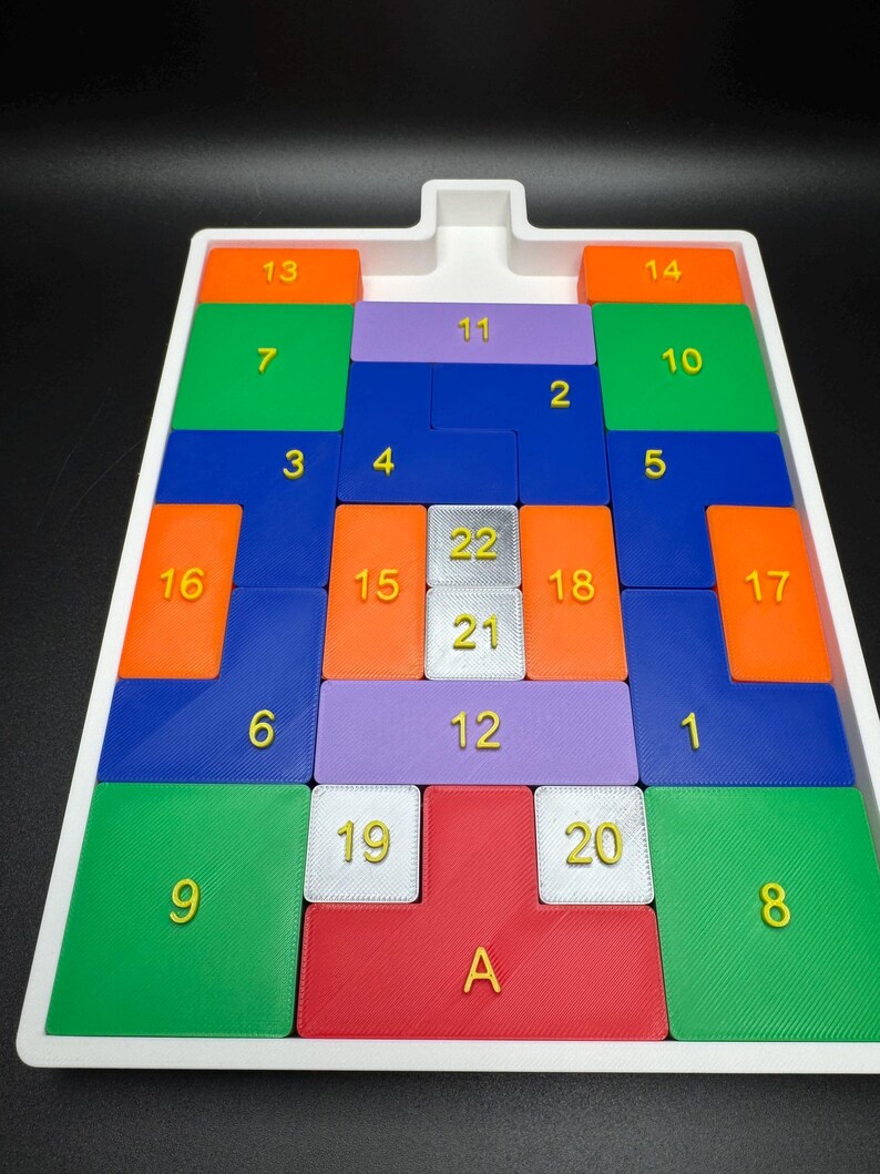 Op de afbeelding: Een witte plastic bak met een puzzelspel. De puzzelstukken zijn gemaakt van verschillende kleuren, waaronder oranje, groen, blauw, paars en rood. De stukken zijn genummerd van 1 tot 22 en A.