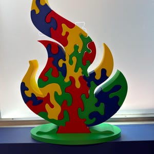 Puede incluir: Un rompecabezas con forma de llama sobre una base ovalada verde. El rompecabezas est&aacute; compuesto por piezas entrelazadas en colores primarios que incluyen rojo, amarillo, azul y verde. El dise&ntilde;o presenta una estructura central vertical.