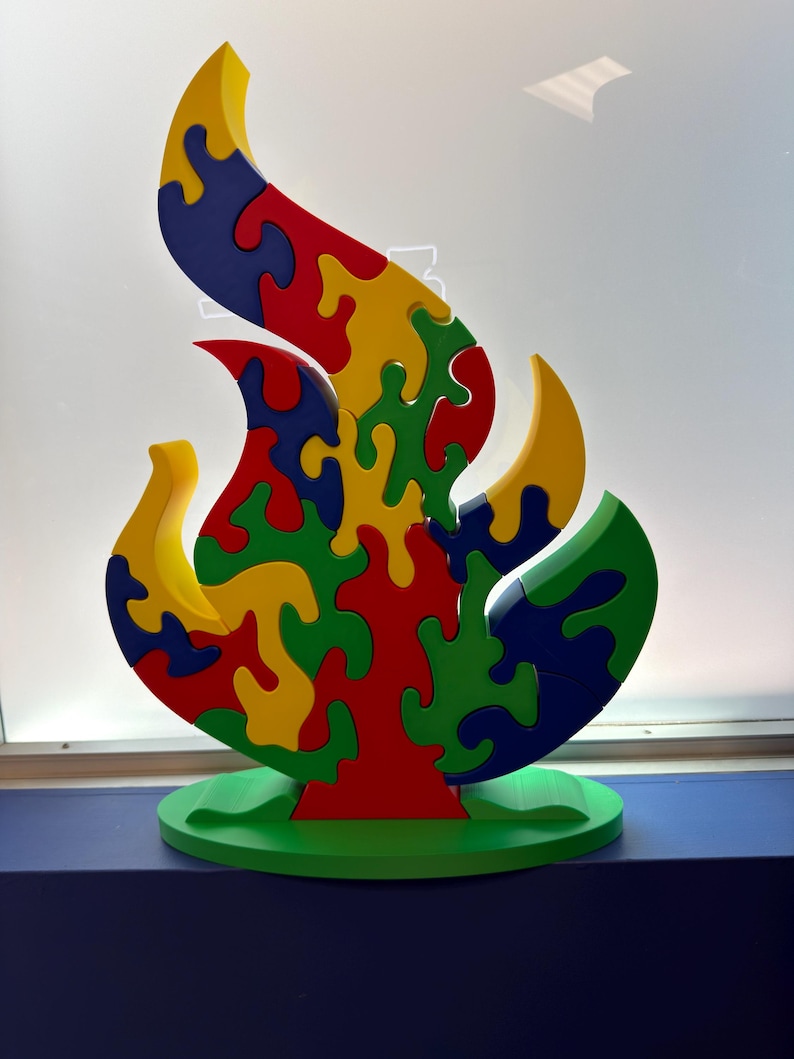 Puede incluir: Un rompecabezas con forma de llama sobre una base ovalada verde. El rompecabezas est&aacute; compuesto por piezas entrelazadas en colores primarios que incluyen rojo, amarillo, azul y verde. El dise&ntilde;o presenta una estructura central vertical.