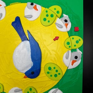 May include: A colorful, 3D-printed bird and egg-themed design on a green and yellow background. The bird is blue and white, with a black eye and orange beak. The eggs are green with yellow spots. The design is on a square, green surface.