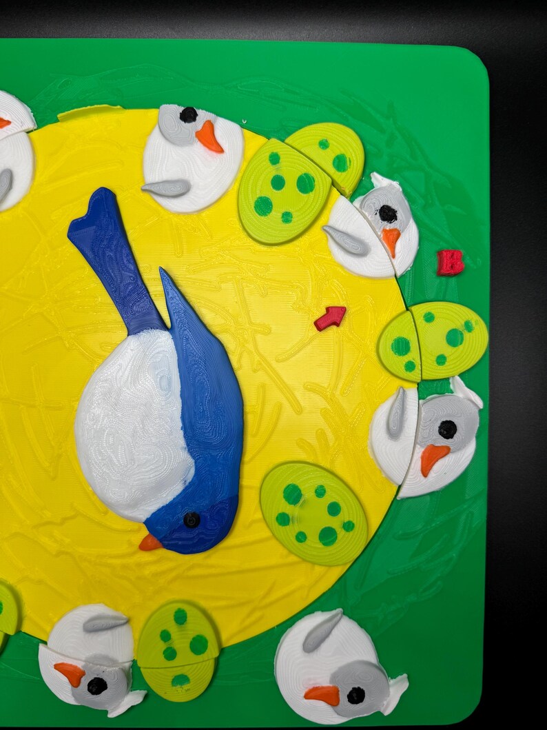 May include: A colorful, 3D-printed bird and egg-themed design on a green and yellow background. The bird is blue and white, with a black eye and orange beak. The eggs are green with yellow spots. The design is on a square, green surface.