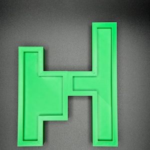 Puede incluir: Una pieza de rompecabezas de pl&aacute;stico verde en forma de letra H. La letra est&aacute; compuesta por cuatro piezas separadas que encajan.