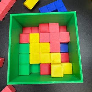 May include: A green square container filled with colorful, geometric blocks. The blocks are red, yellow, green, and blue. Some blocks are outside the container. The blocks are arranged in a Tetris-like pattern.