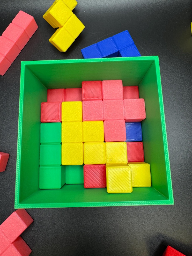 May include: A green square container filled with colorful, geometric blocks. The blocks are red, yellow, green, and blue. Some blocks are outside the container. The blocks are arranged in a Tetris-like pattern.