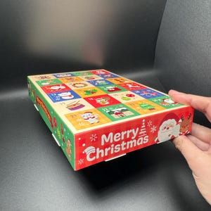 May include: A colorful Christmas advent calendar with numbered doors. The box features a red border with "Merry Christmas" text and festive illustrations of Santa Claus, reindeer, and snowflakes. The top has 24 decorated squares.