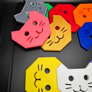May include: A collection of colorful cat-shaped puzzle pieces in various colors, including pink, yellow, and blue. Each piece features a smiling cat face with simple line details. The pieces are arranged on a black surface.