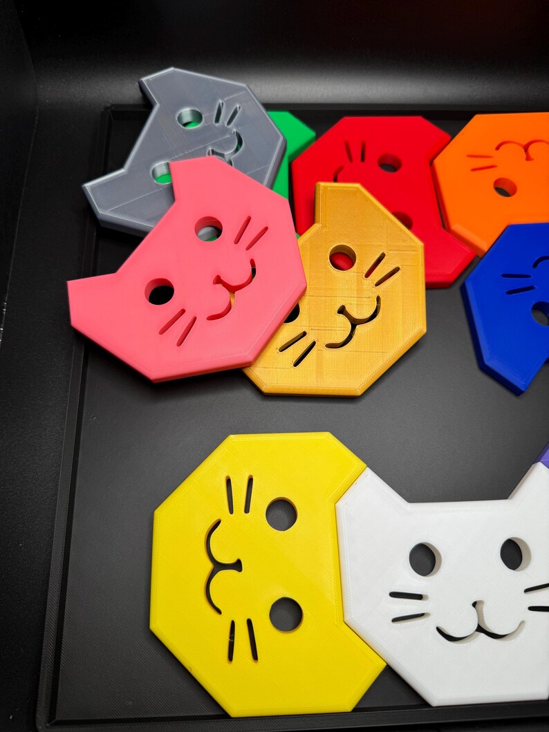 May include: A collection of colorful cat-shaped puzzle pieces in various colors, including pink, yellow, and blue. Each piece features a smiling cat face with simple line details. The pieces are arranged on a black surface.