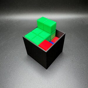 May include: A black square container holds a puzzle with green, red, and blue blocks. The green blocks are stacked on top of the other blocks. The puzzle is on a dark surface.