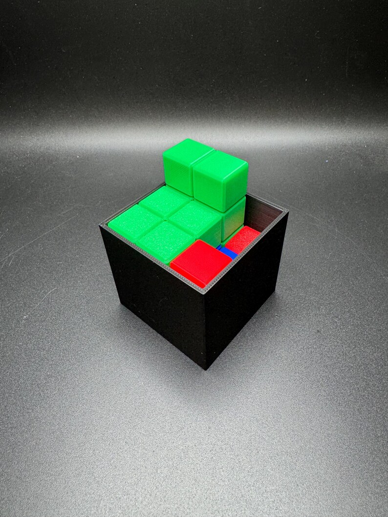 May include: A black square container holds a puzzle with green, red, and blue blocks. The green blocks are stacked on top of the other blocks. The puzzle is on a dark surface.