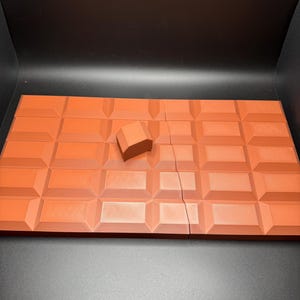May include: A broken chocolate bar with a single piece of chocolate on top. The chocolate is a dark brown colour and has a textured surface.