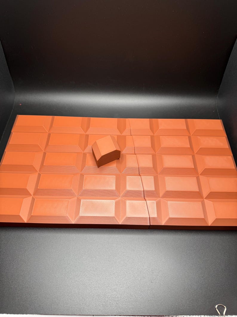 May include: A broken chocolate bar with a single piece of chocolate on top. The chocolate is a dark brown colour and has a textured surface.