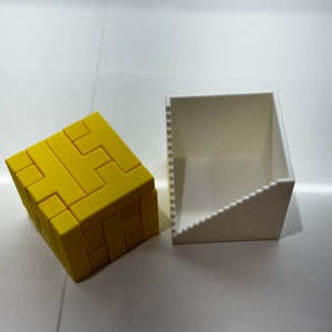 Puede incluir: Un rompecabezas 3D amarillo en forma de cubo, con una caja blanca a su lado. El rompecabezas est&aacute; formado por piezas m&aacute;s peque&ntilde;as que encajan para formar una forma m&aacute;s grande.
