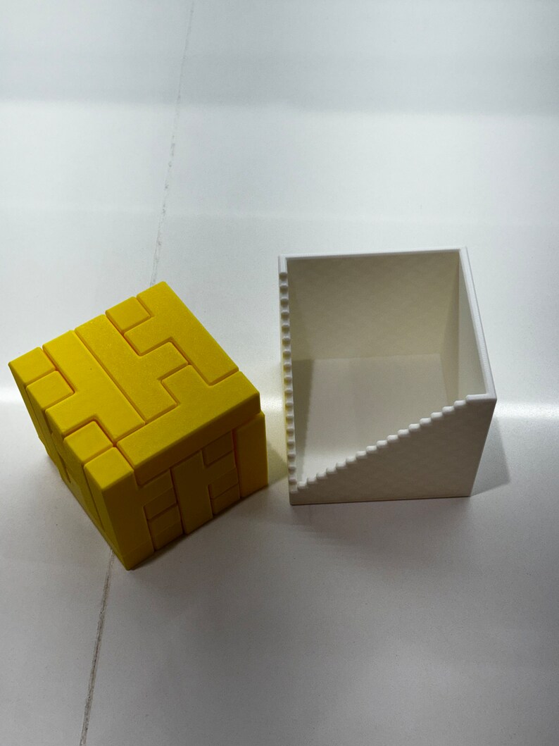 Puede incluir: Un rompecabezas 3D amarillo en forma de cubo, con una caja blanca a su lado. El rompecabezas est&aacute; formado por piezas m&aacute;s peque&ntilde;as que encajan para formar una forma m&aacute;s grande.