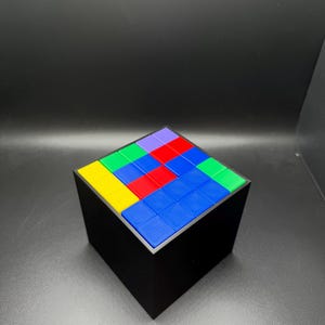May include: A black cube puzzle with a colorful top. The top of the cube is composed of smaller squares in various colors, including blue, red, green, yellow, and purple. The cube is sitting on a dark surface.