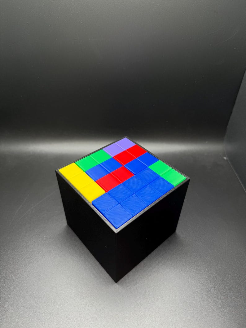 May include: A black cube puzzle with a colorful top. The top of the cube is composed of smaller squares in various colors, including blue, red, green, yellow, and purple. The cube is sitting on a dark surface.
