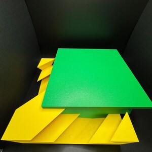 May include: A green square platform sits on top of a yellow geometric base. The base is made of several overlapping triangles, creating a layered effect.