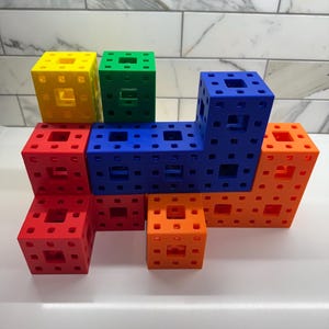 May include: A set of colorful building blocks in red, orange, yellow, green, and blue. The blocks are made of plastic and have a grid pattern on each side. They are arranged in a tower-like structure.