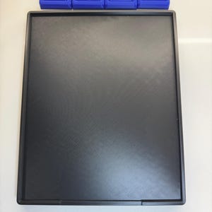 May include: A black rectangular tray with a smooth, textured surface. The tray is made of plastic and has a slightly raised edge.