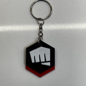 May include: A black, red, and white keychain featuring a hexagonal design with a fist emblem. The keychain has a silver metal ring and chain for attaching to keys or bags. The fist emblem is white on a black background, with a red accent.