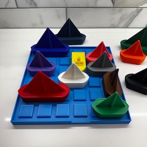 May include: A blue play mat with a grid pattern and various colored toy boats. The boats are made of plastic and include a red, green, blue, white, yellow, purple, orange, brown, and black boat.