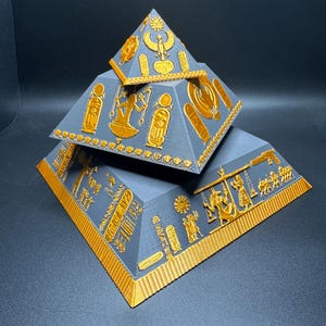 Puede incluir: Modelo de pir&aacute;mide de tres niveles en gris y dorado. Las pir&aacute;mides est&aacute;n apiladas, con la m&aacute;s grande en la base y la m&aacute;s peque&ntilde;a en la parte superior. Los detalles dorados incluyen jerogl&iacute;ficos y otros s&iacute;mbolos egipcios. Las pir&aacute;mides son probablemente de pl&aacute;stico.