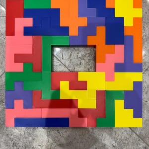 May include: A colorful wooden puzzle with a variety of shapes and colors, including red, orange, yellow, green, blue, and purple. The puzzle is arranged in a square shape with a hole in the center.