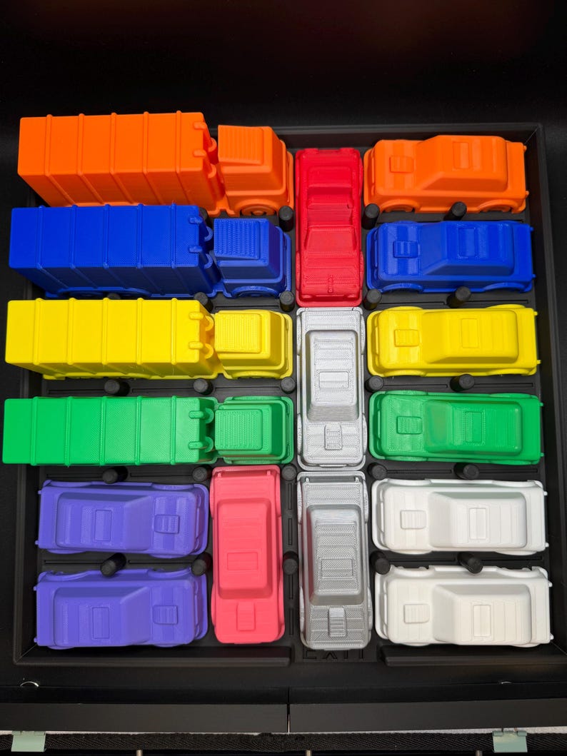 May include: An assortment of brightly coloured toy trucks, including orange, blue, red, yellow, green, purple, grey, and white. The trucks are arranged in a grid, displaying their different designs and sizes. The image is taken from a top-down perspective.