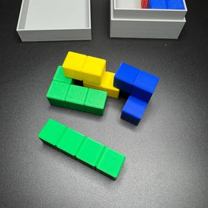 Puede incluir: Un conjunto de bloques de madera de colores en azul, verde y amarillo. Los bloques est&aacute;n dispuestos en una configuraci&oacute;n de rompecabezas. Los bloques est&aacute;n en una caja gris.