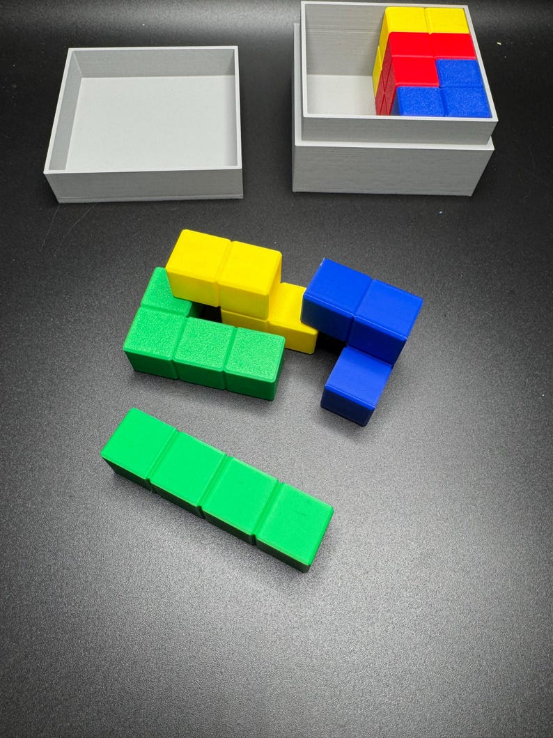 Puede incluir: Un conjunto de bloques de madera de colores en azul, verde y amarillo. Los bloques est&aacute;n dispuestos en una configuraci&oacute;n de rompecabezas. Los bloques est&aacute;n en una caja gris.
