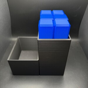 May include: A black 3D printed organizer with a rectangular compartment and a square compartment. The square compartment holds six blue rectangular blocks.