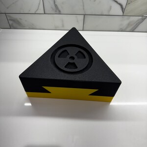 May include: A black triangular box with a yellow stripe and a raised radiation symbol on the top.