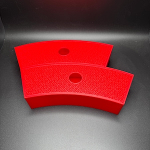 May include: Two bright red, curved plastic trays with a textured surface. Each tray has a circular hole in the center. The trays are stacked on top of each other against a dark background.