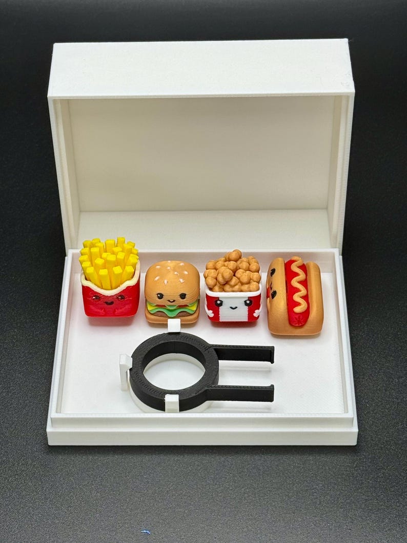 May include: A white box with a black ring inside. The box contains four small, colorful figurines of fast food items: french fries, a hamburger, chicken nuggets, and a hot dog. Each figurine has a cute cartoon face.