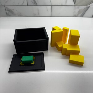 Pu&ograve; includere: Una scatola nera con un pulsante verde e un coperchio. La scatola &egrave; aperta e ci sono blocchi gialli sparsi intorno.