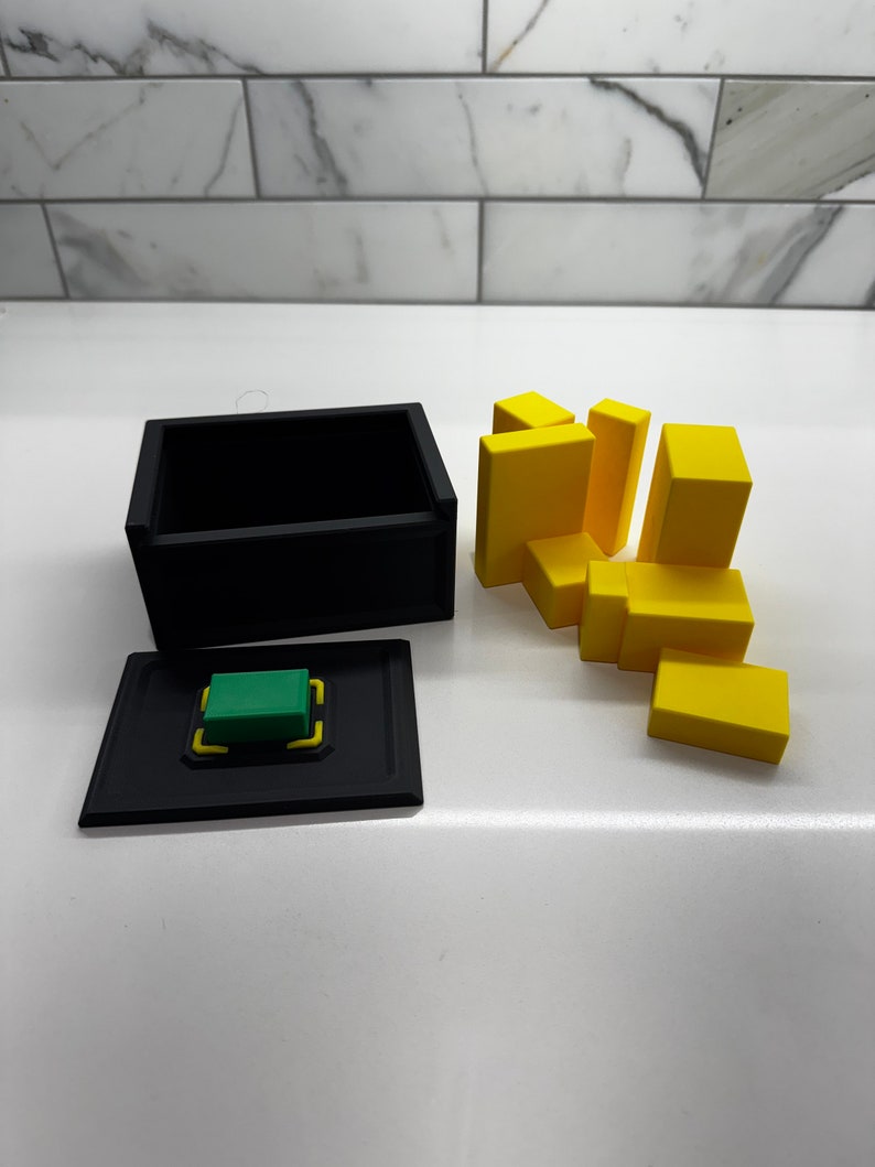 Pu&ograve; includere: Una scatola nera con un pulsante verde e un coperchio. La scatola &egrave; aperta e ci sono blocchi gialli sparsi intorno.