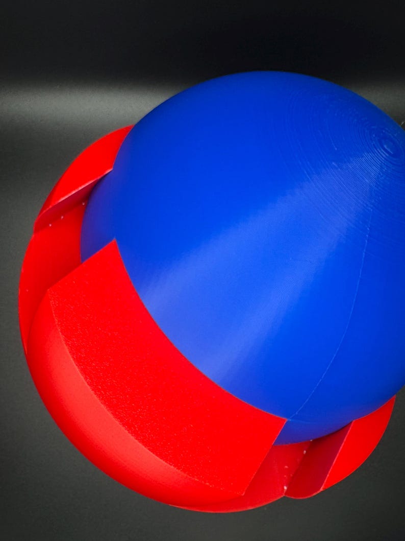 Puede incluir: Una esfera impresa en 3D con la parte superior azul y la inferior roja. La esfera est&aacute; hecha de dos mitades que encajan.