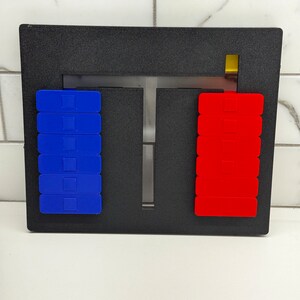 May include: A black frame with a T-shaped opening in the center. There are two stacks of blocks, one blue and one red, on either side of the opening. A small yellow block is visible at the top of the frame.