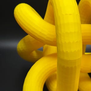 May include: A yellow 3D printed knot sculpture. The knot is made of multiple intertwined loops, creating a complex and intricate design.