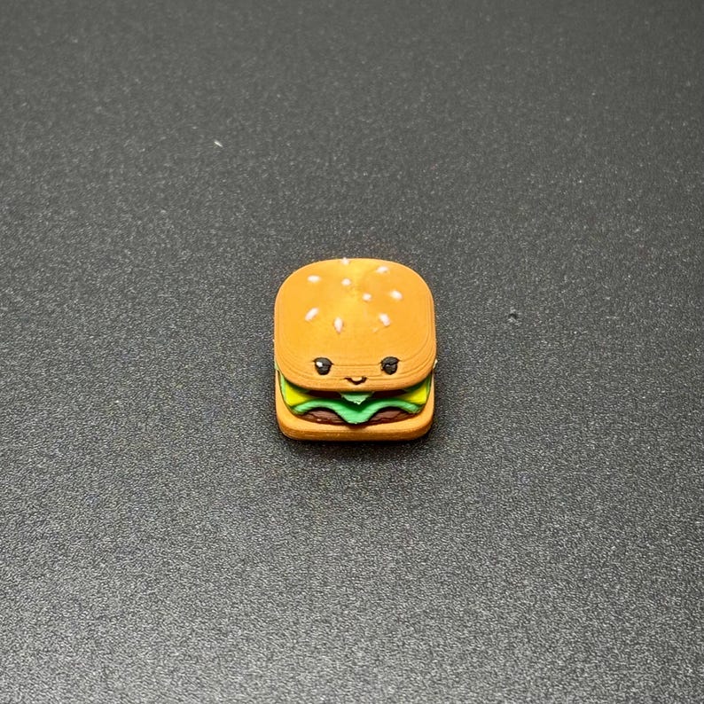 May include: A small, cartoon-style hamburger with a sesame seed bun, lettuce, and tomato. The hamburger has a happy face with black eyes and a smile.