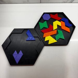 Op de afbeelding: Een zwarte zeshoekige puzzelbak met een paars hartvormig stuk in de onderste bak en een verscheidenheid aan gekleurde driehoekige stukken in de bovenste bak.