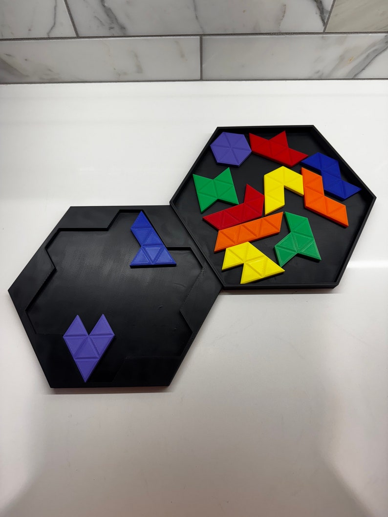 Op de afbeelding: Een zwarte zeshoekige puzzelbak met een paars hartvormig stuk in de onderste bak en een verscheidenheid aan gekleurde driehoekige stukken in de bovenste bak.
