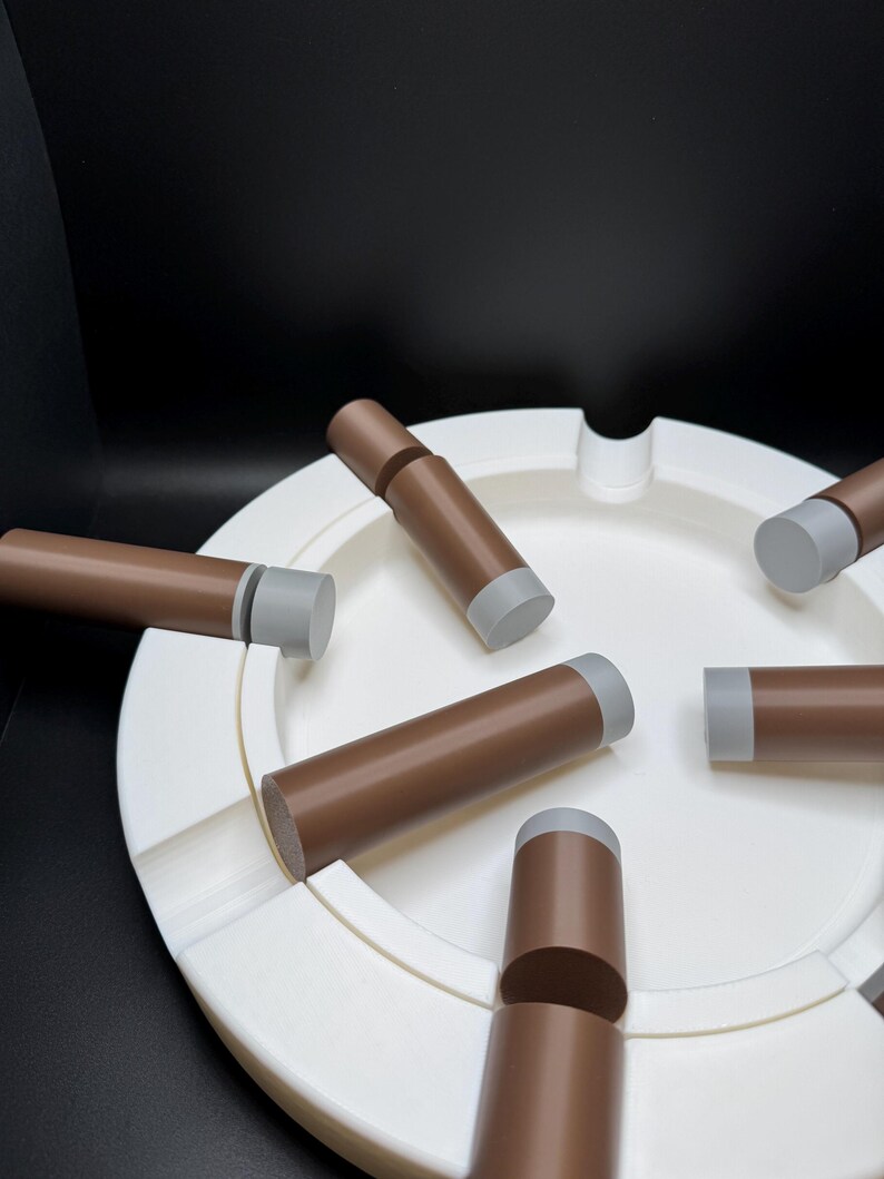 K&ouml;nnte beinhalten: Ein wei&szlig;er Aschenbecher mit kreisf&ouml;rmigem Design, der mehrere braune und graue zigarrenf&ouml;rmige Objekte aufweist. Die Zigarren sind um den Aschenbecher herum angeordnet und bilden eine ansprechende Anordnung. Der Hintergrund ist einfarbig schwarz.