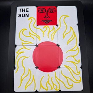 May include: A puzzle featuring the sun tarot card design. The puzzle has a black frame and is composed of white squares with yellow flames and a red sun in the center. The text "THE SUN" is visible on one of the squares.