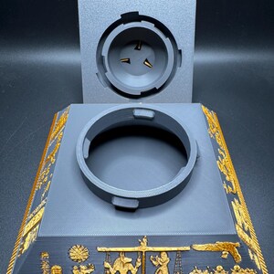 Puede incluir: Un soporte gris impreso en 3D con una abertura circular y una tapa separada. El soporte presenta dise&ntilde;os jerogl&iacute;ficos egipcios dorados a lo largo de los bordes y la base, con un relieve detallado de figuras y s&iacute;mbolos.